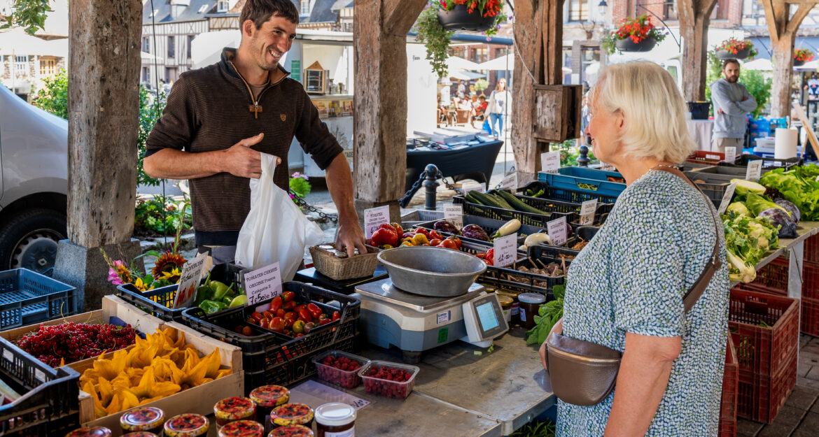 Commerçant souriant vendant des légumes et fruits à une cliente sur le marché de Lyons-la-Forêt, sous une halle en bois.