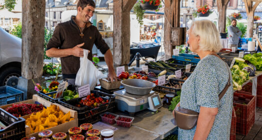 Commerçant souriant vendant des légumes et fruits à une cliente sur le marché de Lyons-la-Forêt, sous une halle en bois.