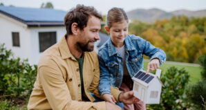 Un homme et une enfant manipulent une maquette de maison avec panneaux solaires photovoltaïques, avec une maison réelle équipée en arrière-plan.